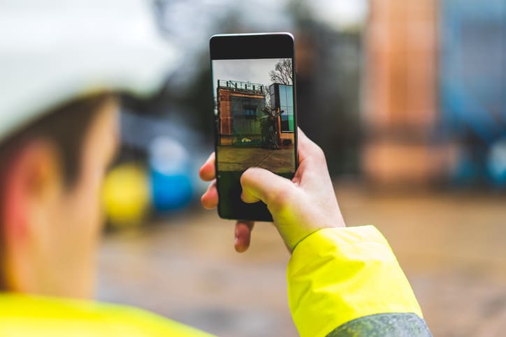 Architects holding a smartphone on construction site - young construction worker is using mobile phone on site - Construction worker with building plans and cellphone - Focus on mobile. warm filter
